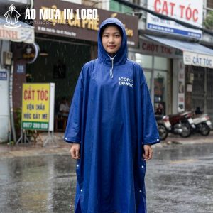 Một bạn nữ mặc áo mưa màu xanh, đội mũ trùm đầu, đứng trên vỉa hè ẩm ướt với nụ cười thân thiện.