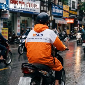 Góc nhìn từ phía sau một người đang lái xe máy, cho thấy rõ logo SHB và thông điệp in sắc nét trên lưng áo mưa.